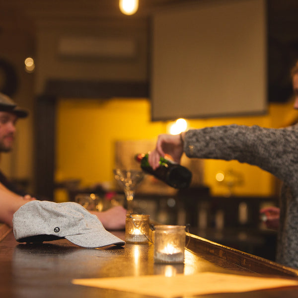 Velo/City Cap - Speckled Hemp Wool Cap lying on a bar-top while drinks are poured in the background.