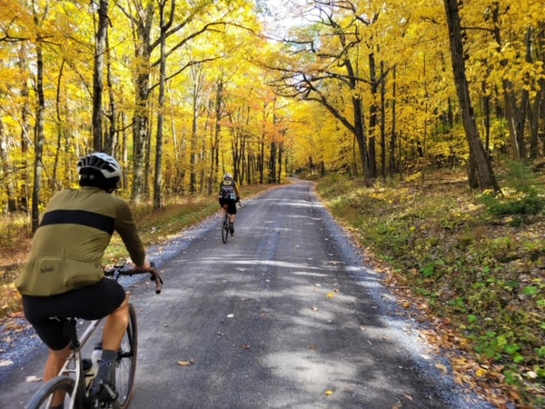 Two cyclists on a country road in the fall, wearing the merino wool long sleeve jersey.